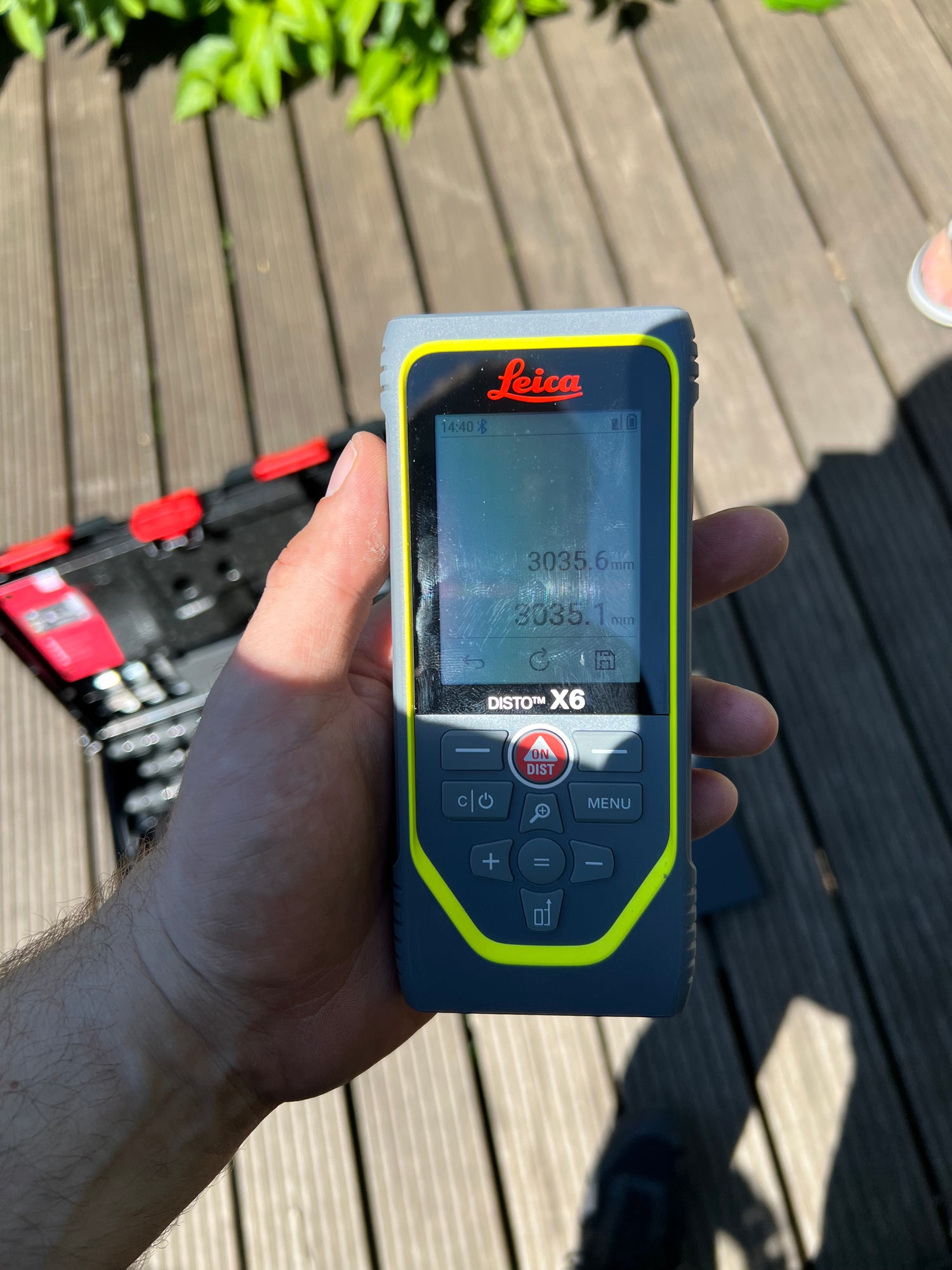 Das Bild zeigt eine Hand, welche ein Laserdistanzmessgerät von Leica hält. Im Hintergrund ist verschwommene eine Terrasse mit Holzdielen sowie der Werkzeugkoffer, in dem vermutlich das Messgerät, war zu sehen.