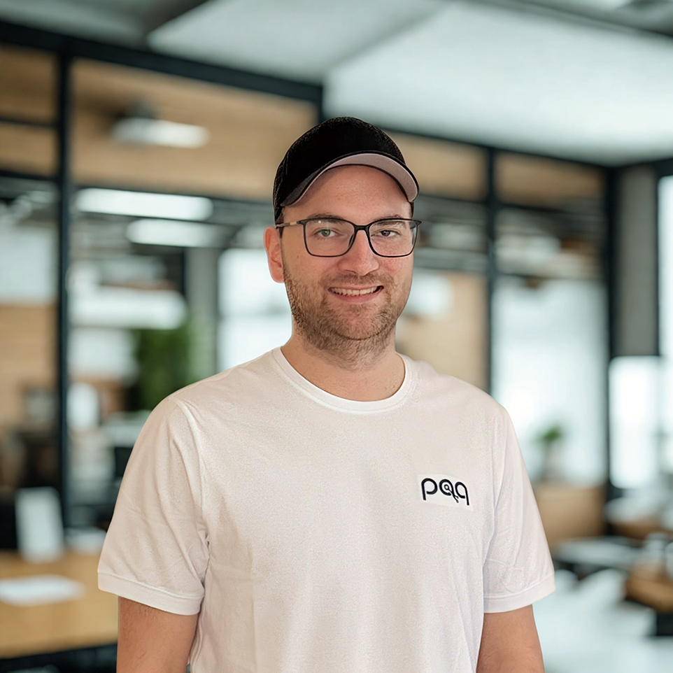 Das Bild zeigt ein Portrait eine Person mit einer schwarzen Kappe, Brille und weißem T-Shirt. Im Hintergrund ist verschwommen eine moderne Bürowand zu sehen