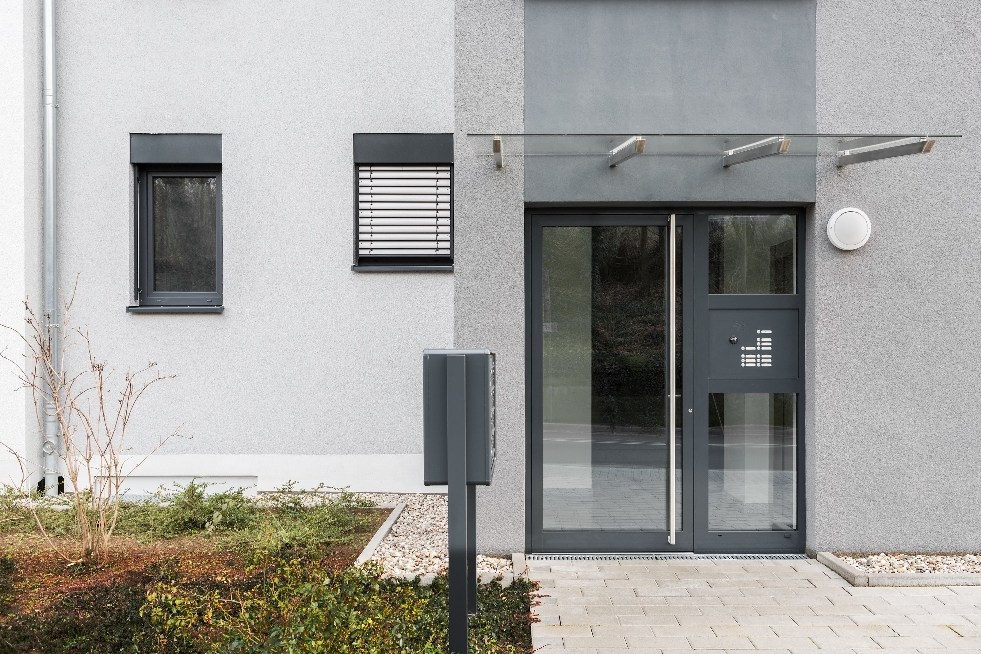 Auf dem Bild ist ein anthrazitfarbene Haustür in einem modernen Gebäude zu sehen. Darüber ist ein Edelstahl/Glas Vordach angebracht. Im Vordergrund steht eine Briefkastenstele