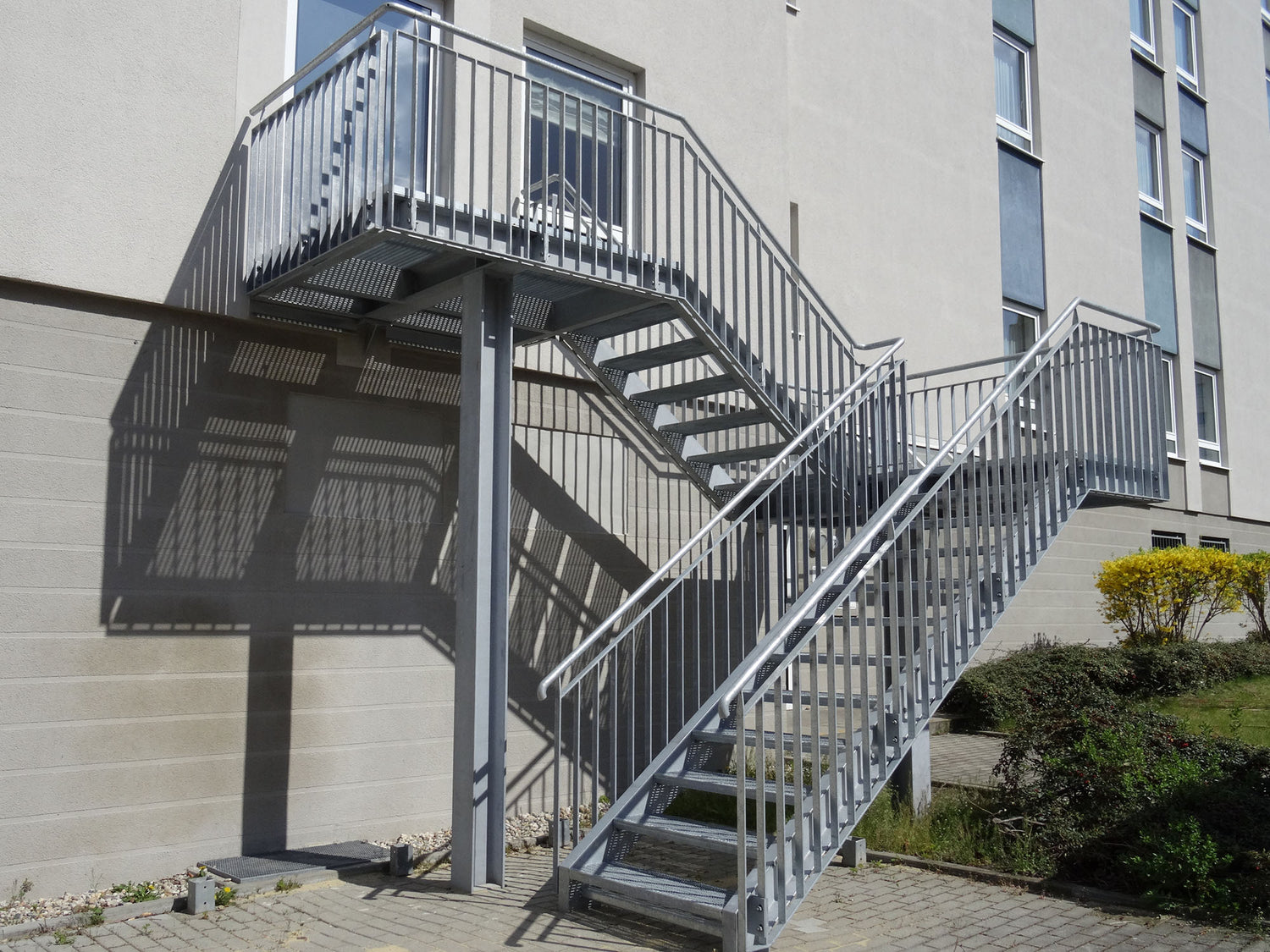 Das Bild zeigt eine Treppe als verzinkte Stahlkonstruktion vor einem modernen mehrstöckigem Gebäude. Das Geländer der Treppe ist als Flachstahlgeländer mit Edelstahlhandlauf ausgeführt. Der untergrund ist gepflastert.