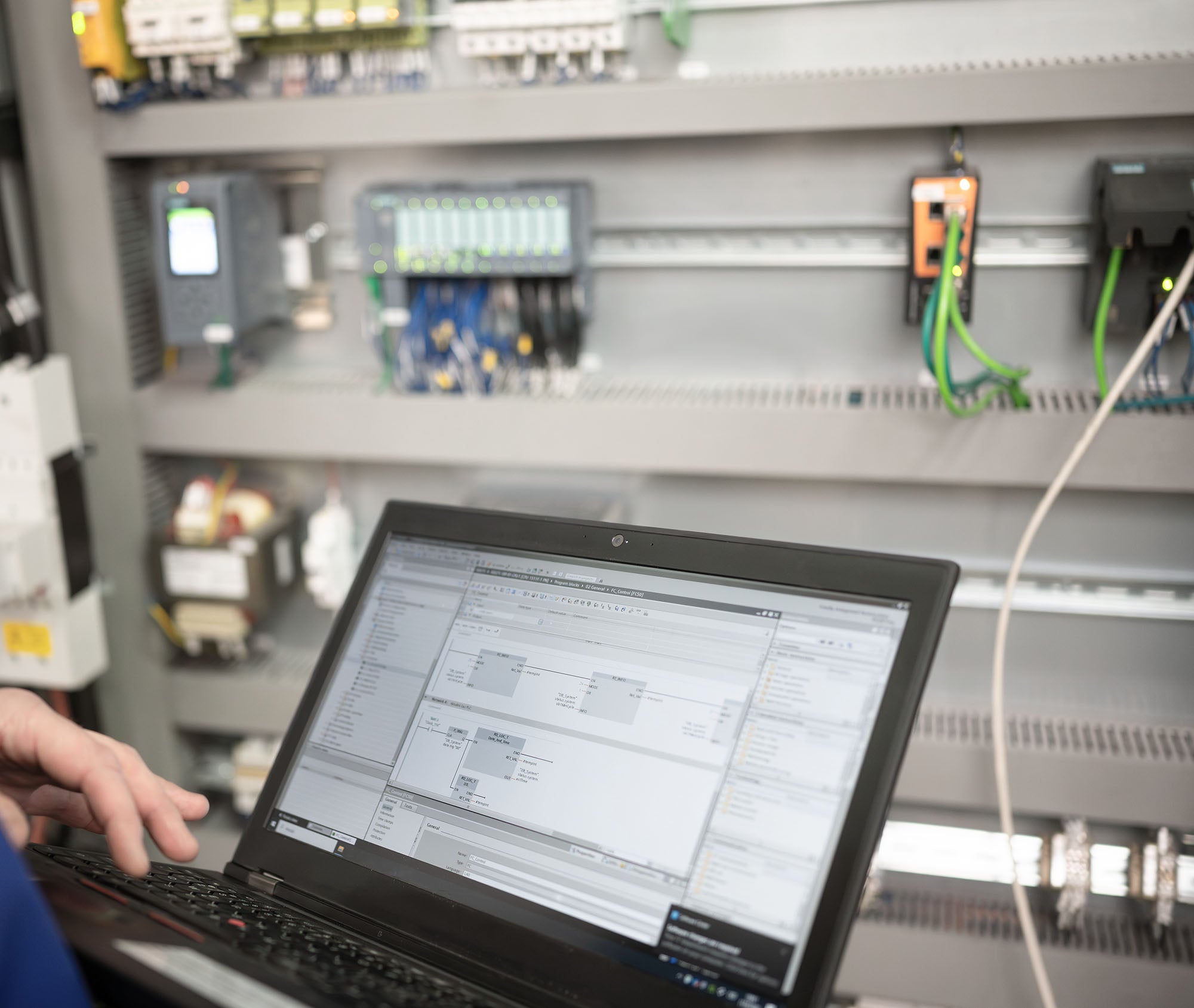 Auf dem Bild ist ein Laptop zu sehen, welcher vor einem Schaltschrank steht. Auf dem Bildschirm sind Programme zu sehen, mit welchen eine SPS Steuerung programmiert wird.
